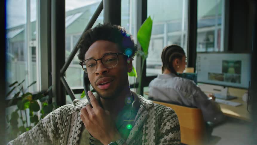 Man talking on the phone in an office and a woman behind facing a screen working