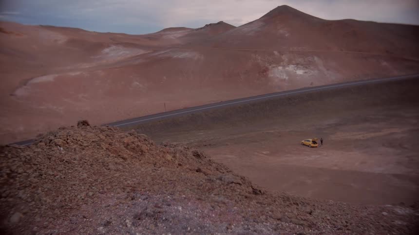 Top view of a cliff in desert mountains with a person next to a yellow vehicle below