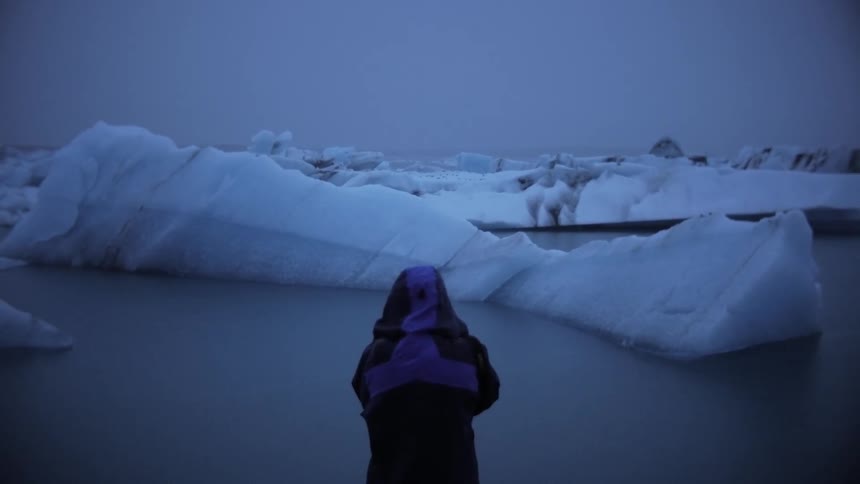 View of a person wearing a hood in front of the arctic with icebergs across the ocean on a dark sky day