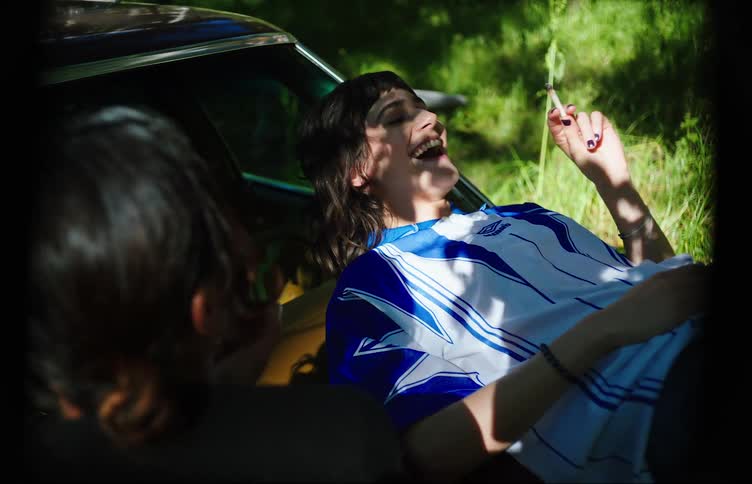 Two young women on the hood of a car with one of them talking and smoking a cigarette