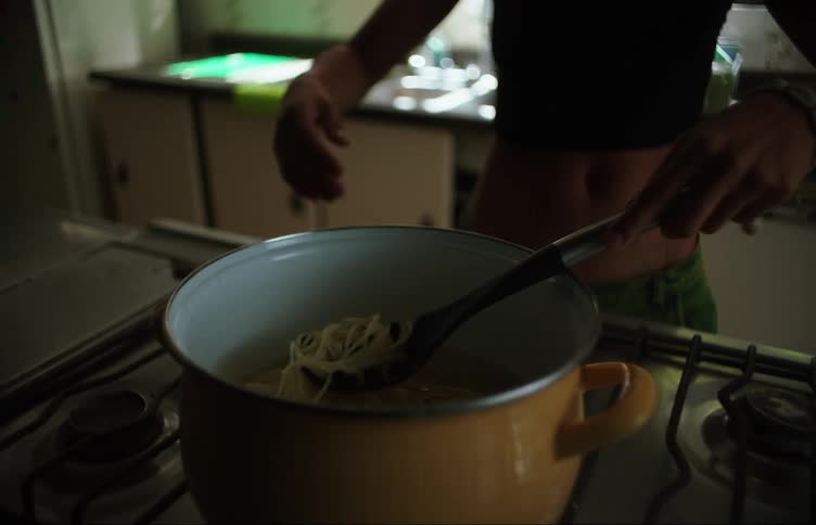 Woman sitting at the kitchen sink bored getting up and checking the pasta in a pan taking out some and throwing it at the wall