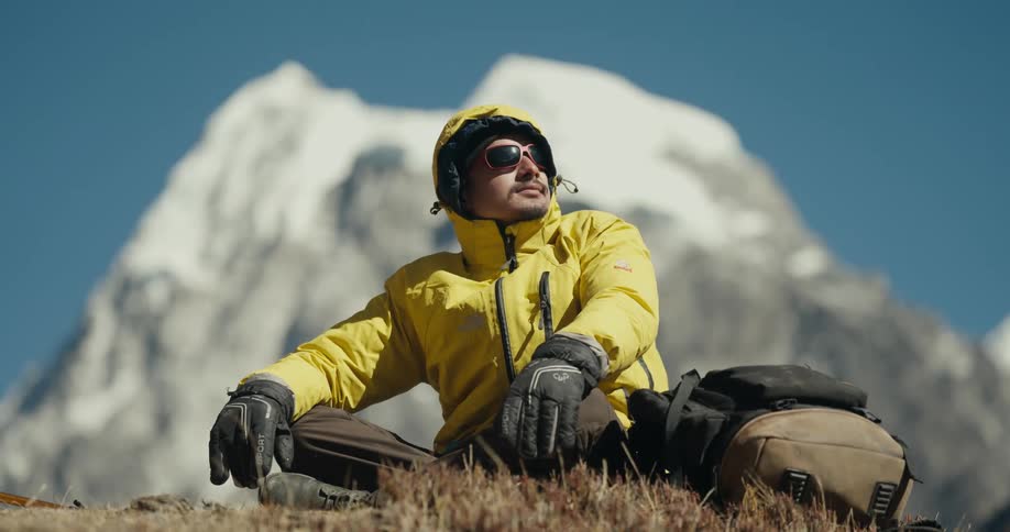 A man wearing glasses and a yellow jacket sits in an icy region and behind him is a snowcovered mountain peak
