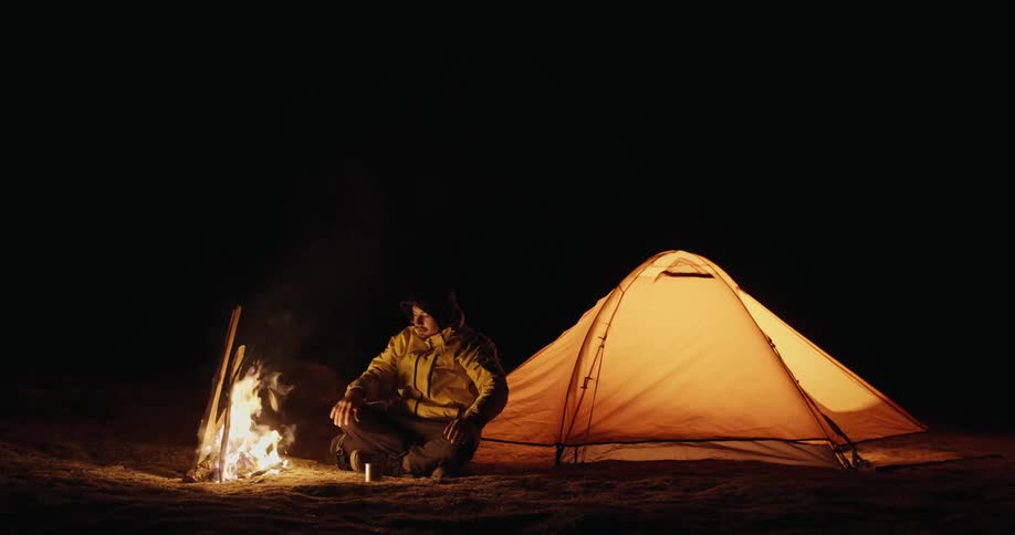 Man sitting in front of a fire warming himself with his tent pitched right behind in a natural environment at night