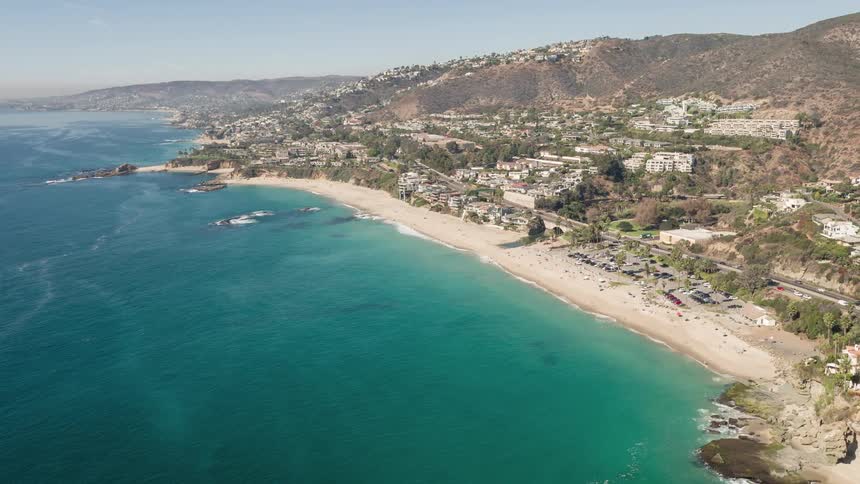An aerial view of a beach with crystal clear water and some houses on land next to a highway with cars passing by