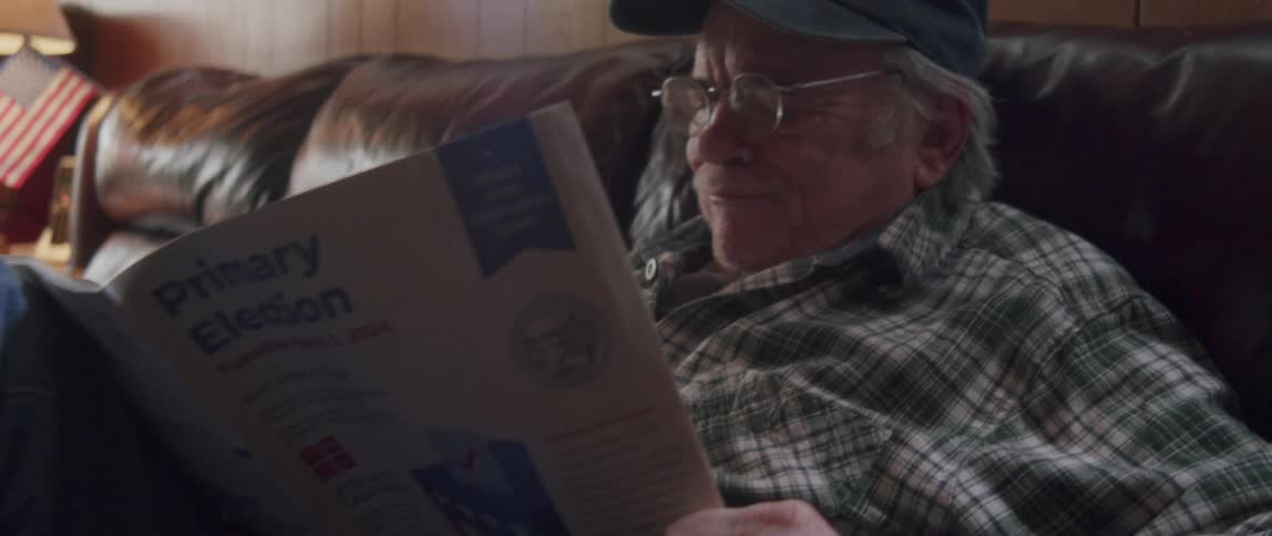 An elderly man wearing a cap and glasses is sitting on a sofa and reading a magazine
