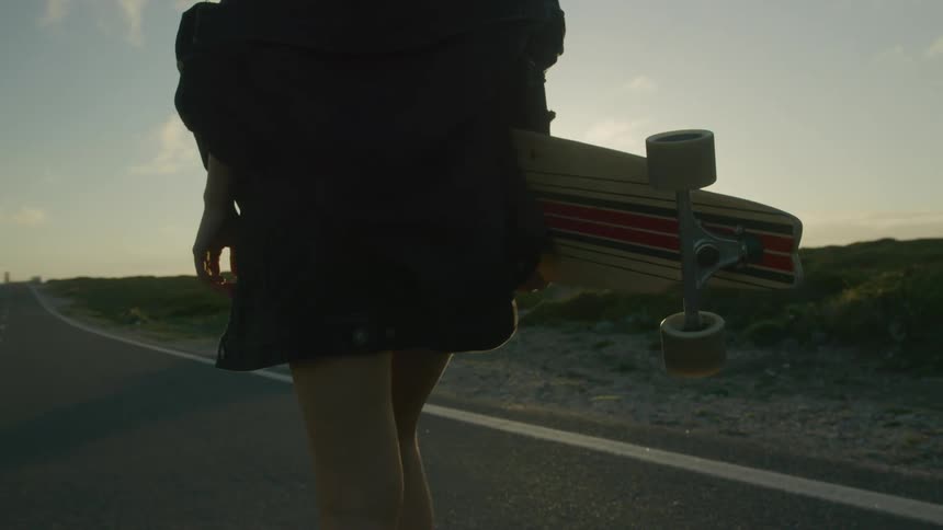 Woman carrying skateboard walks along highway as sun hovers on the horizon