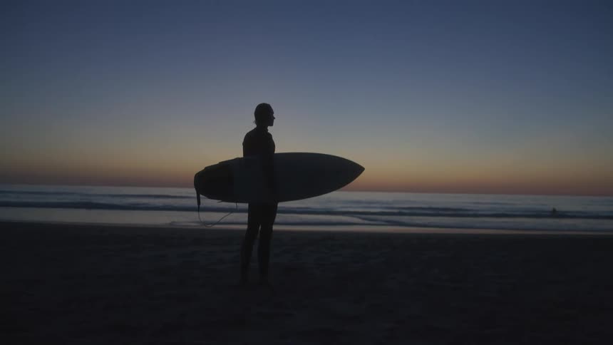 A surfer stands while holding his board on a sandy beach next to the ocean
