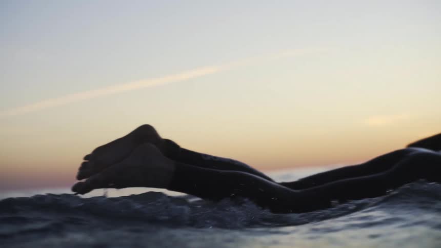 Surfer wearing a wet suit paddles across ocean on surfboard under a red tinted sky