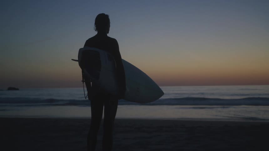 A male surfer stands on the beach and looks out at the ocean while holding his board