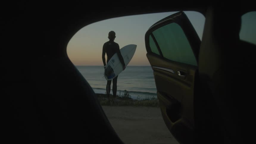 A view from inside a car of a surfer who is standing looking out to sea with a surfboard in his arms