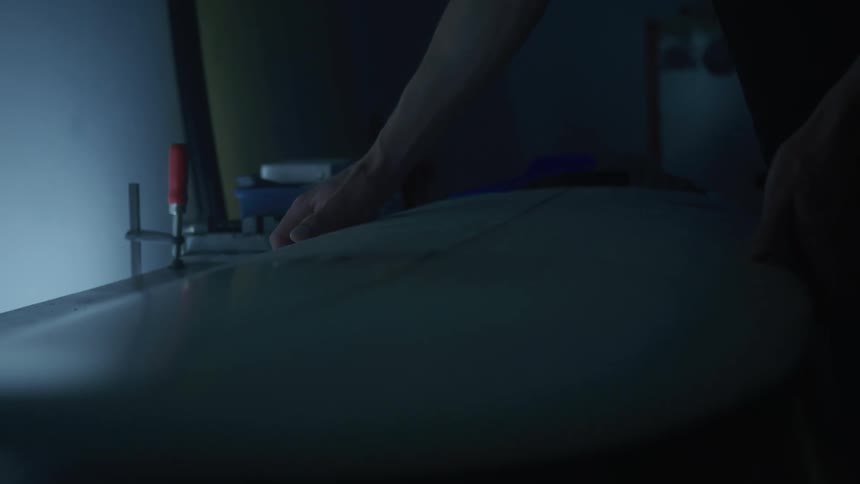 A pair of hands carefully putting down a surfboard on a work bench