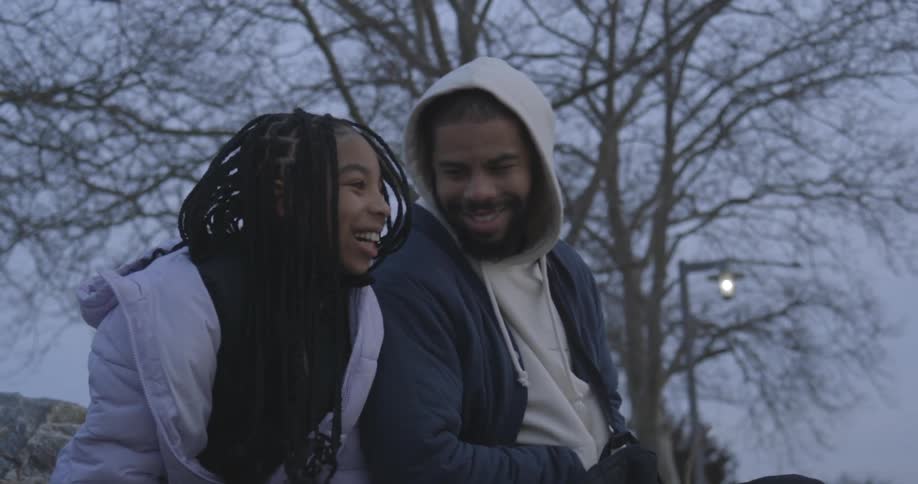 A couple of a man and a woman who are next to each other are sitting and talking while laughing and behind there is a dry tree