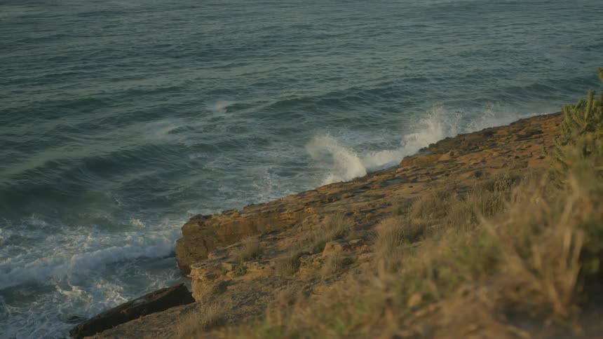 Waves foam as they hit the rocks on shore at the bottom of a grassy slope