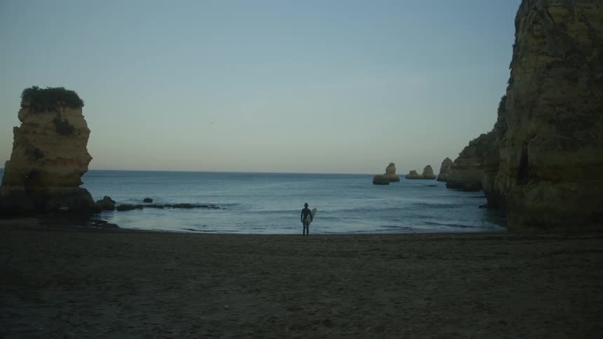 A surfer stands on the shore of a beach at dusk