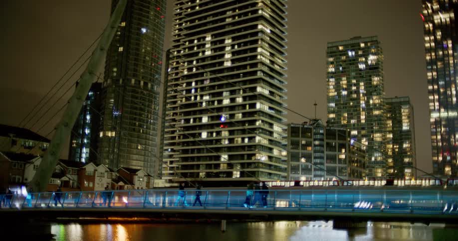 Several people walking along a walkway that goes over water in a city at night