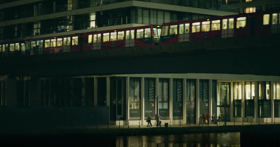 People walking alongside water as a train passes on an overhad bridge