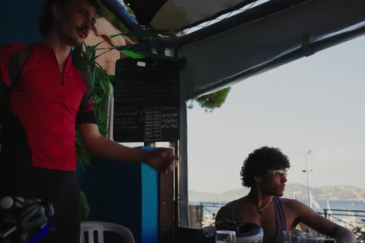 A man in a red shirt is standing and greeting some people who are sitting and then he also sits down