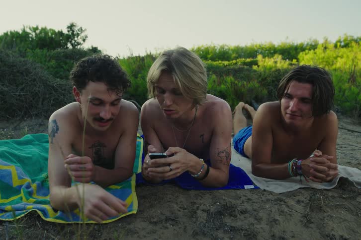 Men sunbathing on beach talk and smile about content on smartphone