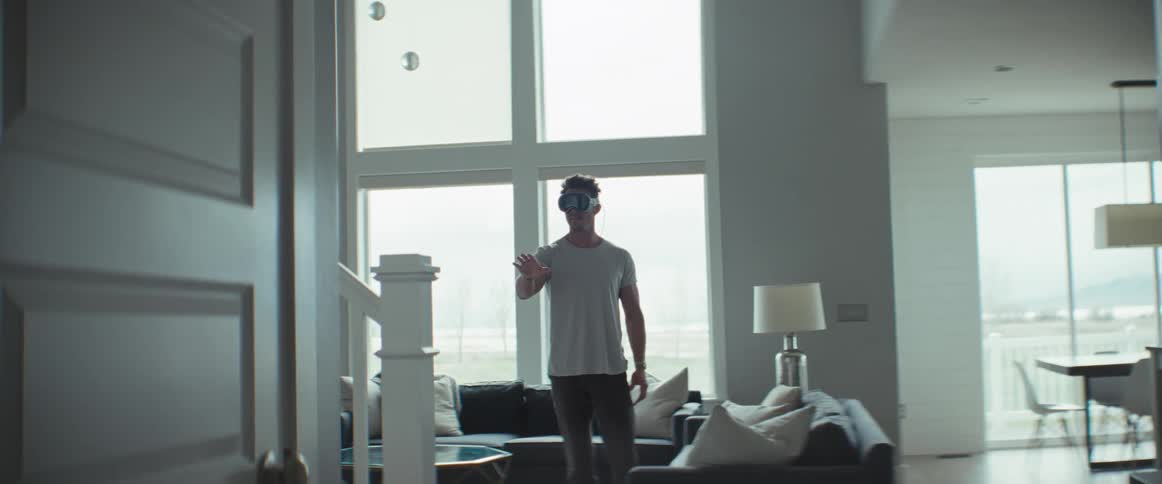 An adult man is standing in an allwhite living room and is wearing virtual reality glasses with his arm raised in front of him
