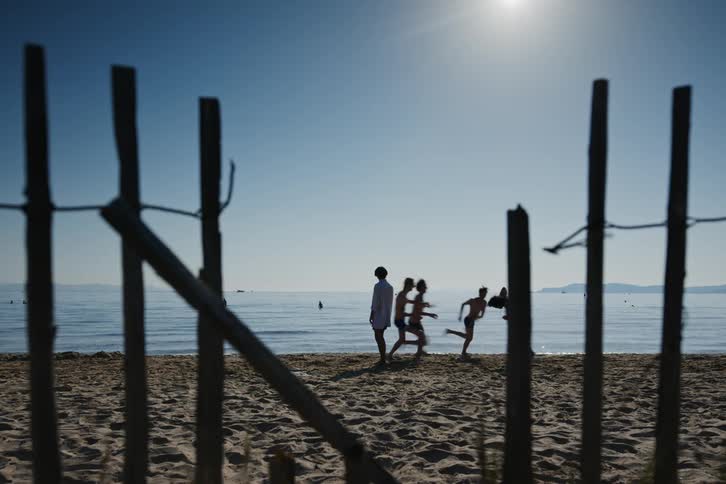 Two people on their feet are looking at each other in front of the beach and then a group of friends run towards one of them and they play