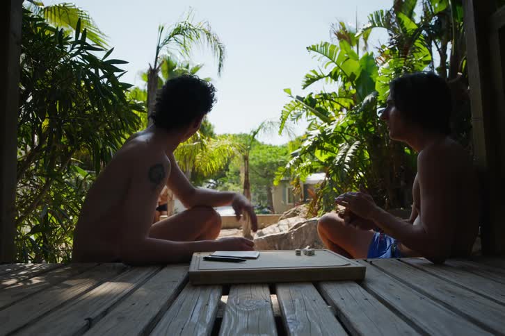 Two friends sitting on a wooden deck in a tropical environment having fun while a young man plays a board game