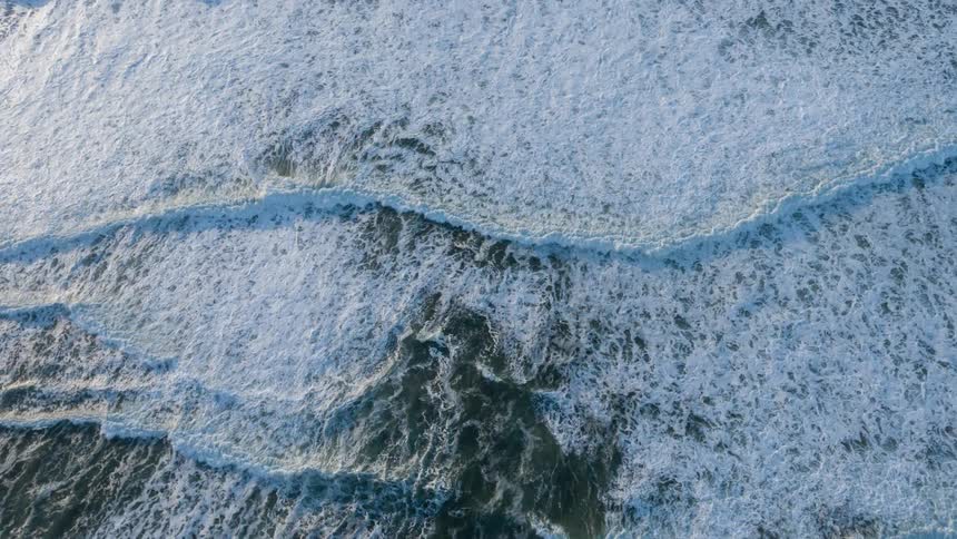 Aerial panorama of a foamy sea with waves passing by and the suns reflection