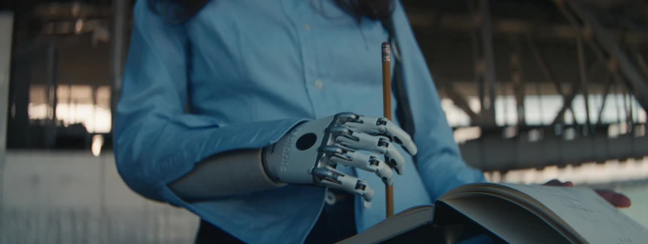 A woman with a mechanical prosthetic in place of a hand and arm uses it to run a pencil over a notebook