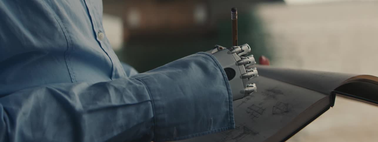 A person in a blue longsleeved tshirt with a robotic hand holding a pencil drawing on a sheet of paper
