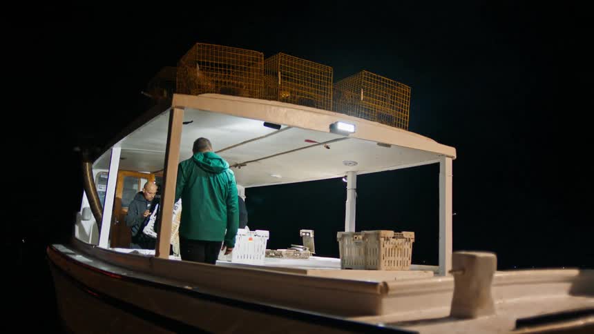 A man in a blue jacket is riding on a boat with two other men who are packing some crates and it is nighttime