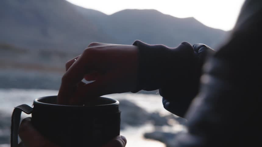 A person in the mountains using a spoon to stir a cup of hot liquid