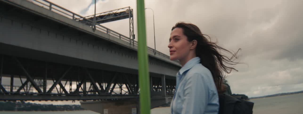 A woman smiling and walking along a river bank next to a large bridge