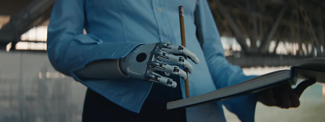 A woman using a prosthetic mechanical hand to write on a notebook with a pencil