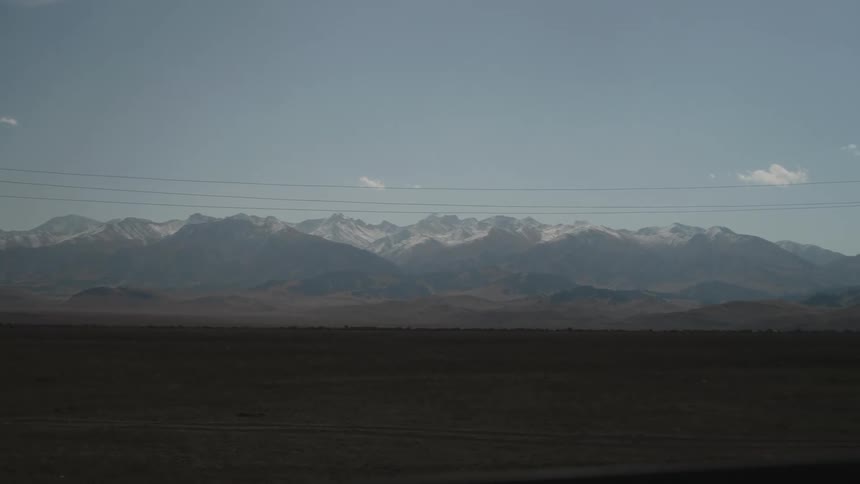 A vast dark expanse of land in the foreground leading to a distant mountain range with snowcapped peaks under a pale blue sky with power lines stretching across the horizon