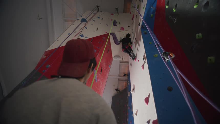 A man watches a woman climbing a wall in a gym