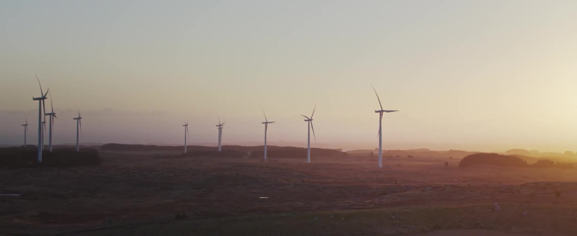A large farm with large windmills that have their propellers rotating and the sun is setting on the horizon making a strong orange light in the sky