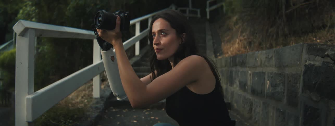 A woman putting her knee to the ground to look through her camera and take a photo as she holds it with both hands including her mechanical prosthesis