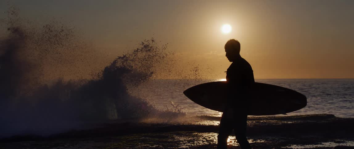 A person with a surfboard walks by crashing waves on a beach
