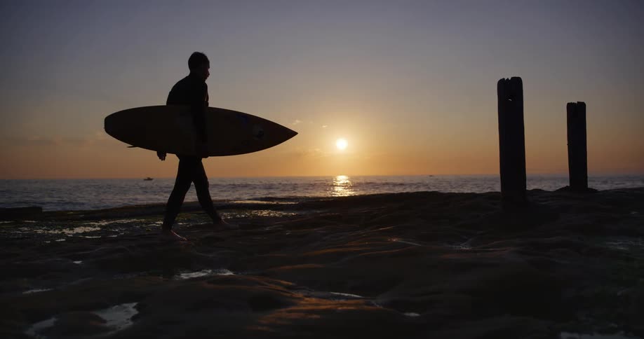 Man walking on the coast with his surfboard near the sea and with the sunrise on the horizon