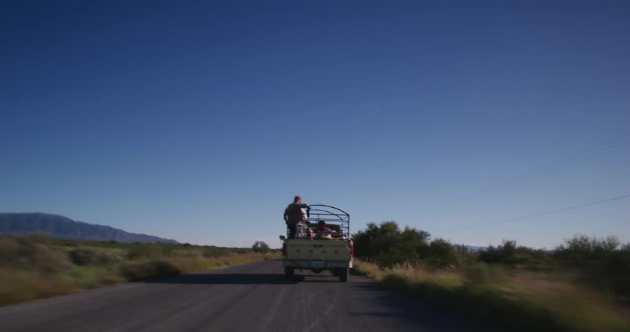 A pick up truck with passengers on the back drives on a rura road