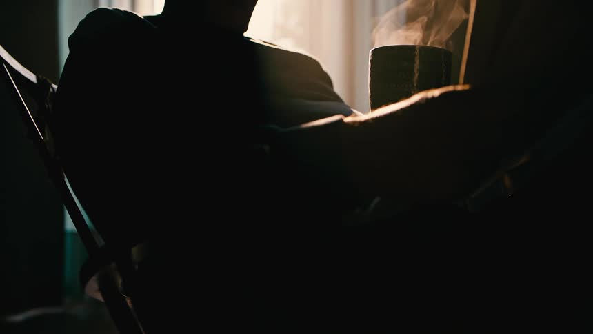 Silhouette of an elderly man sitting a dark room reading and holding a steaming cup