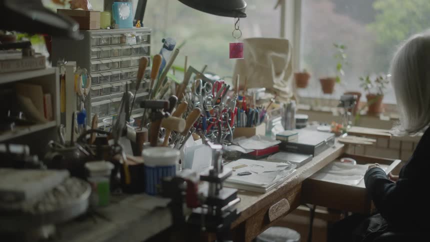 An elderly woman sits down at a cluttered desk and opens a container