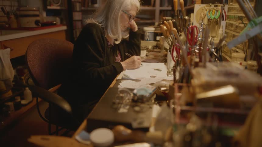 An artist sketching at a cluttered desk filled with art supplies in a cozy room