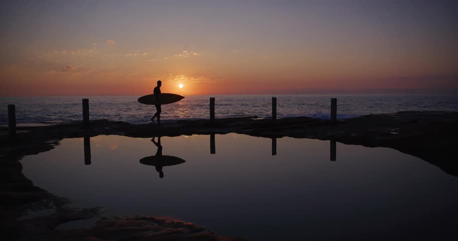Silhouette of a surfer walking next to a tide pool next to the beach at sunset