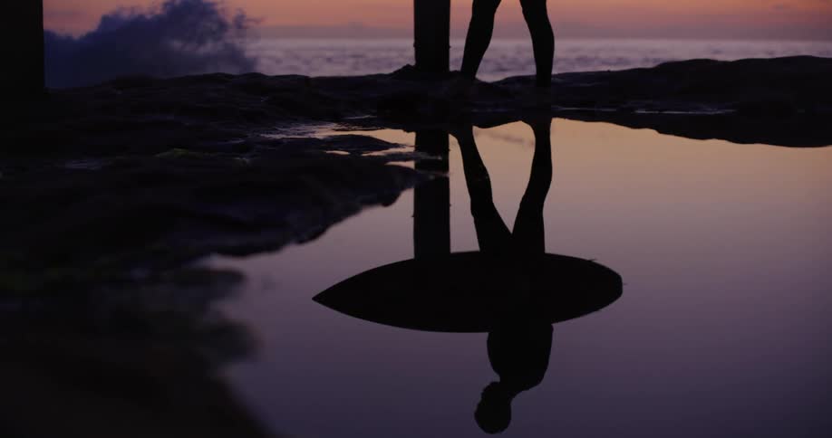 Reflection of a man with a surfboard walking on a rock by the sea on a morning with a purple sky