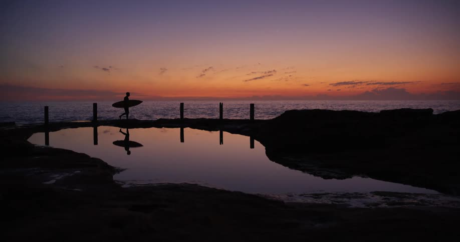 A man with a surfboard walks across the beach at dusk