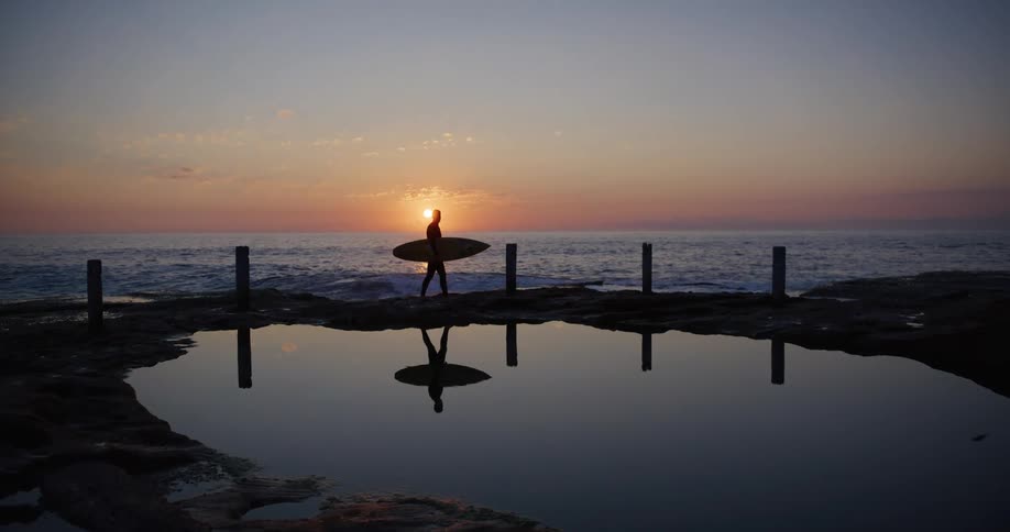 A surfer walking on a beach at sunset with wooden poles and a beach lake to his side