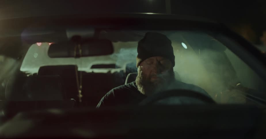 Man with his eyes closed inside a car at night and smoke inside begins to dissipate