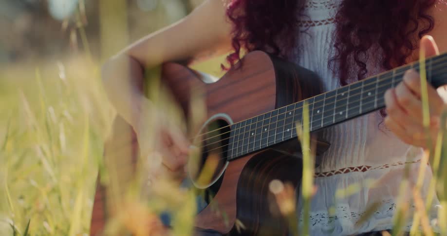 A female plays a guitar while sitting in a meadow on a sunny day