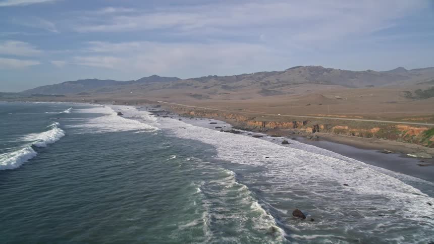 Aerial view of the sea reaching the rocky coast with mountains on the horizon