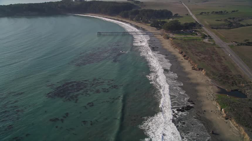 Aerial view of a sea with long pier with waves crashing on the shore with lush vegetation and hills adorn the background